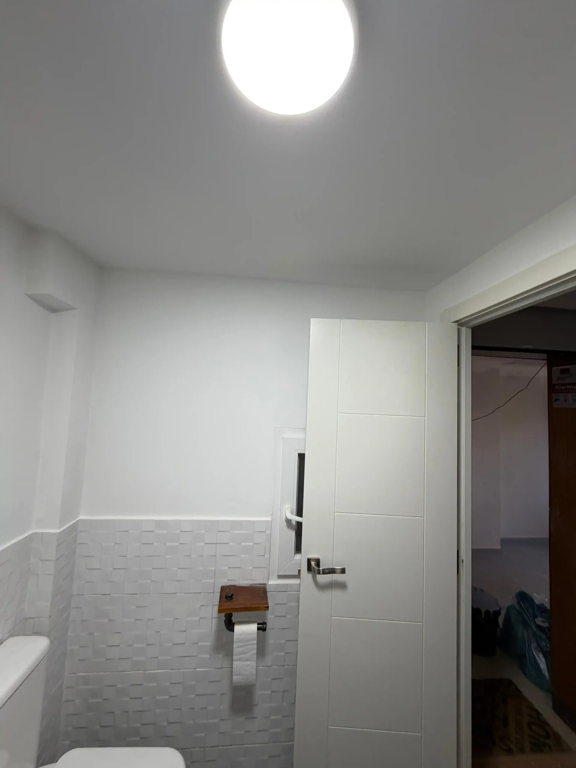 Baño pequeño de color blanco con paredes inferiores alicatadas, puerta abierta y lámpara de techo.