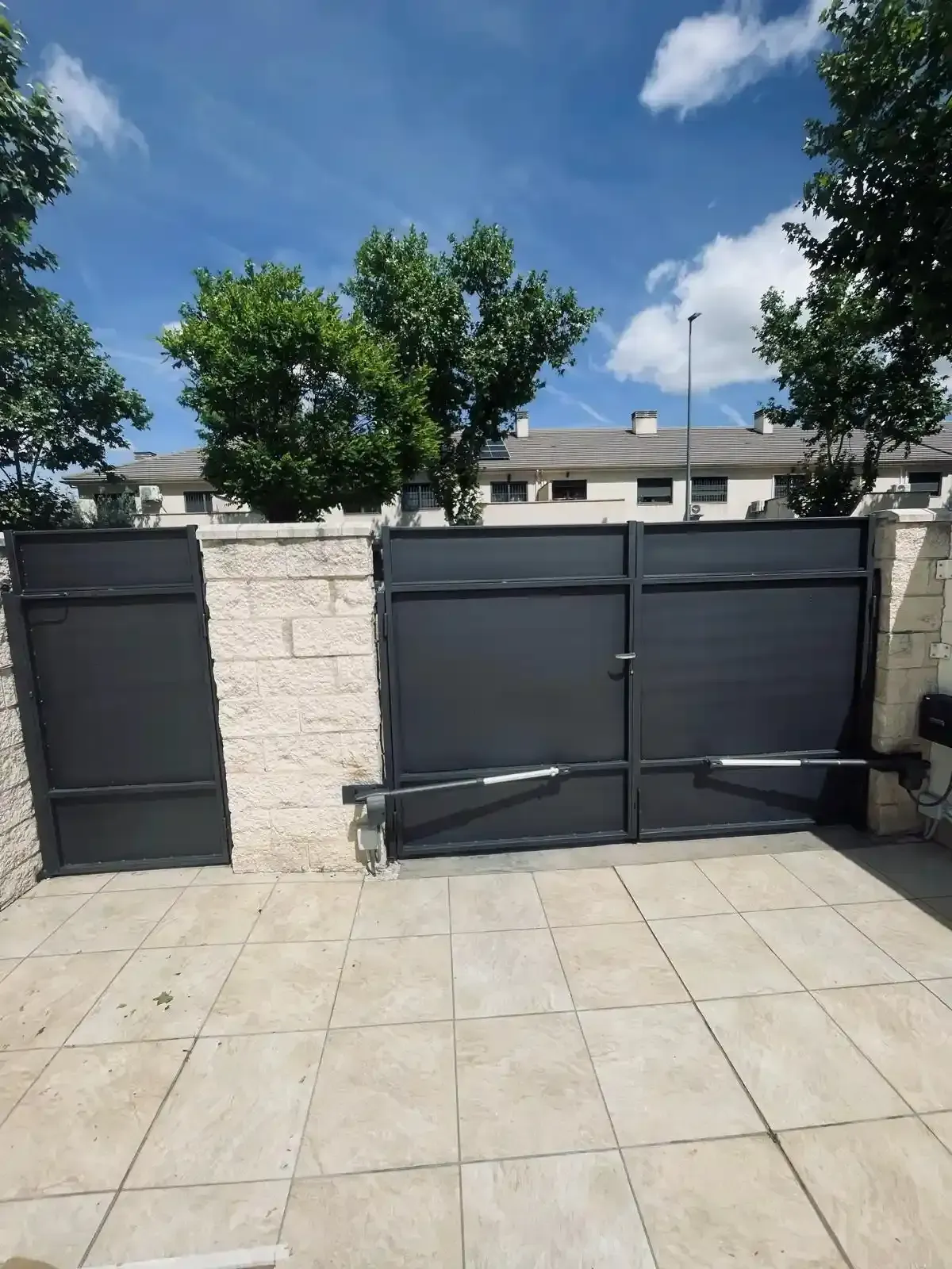 Patio con pavimento beige, pared blanca y verjas metálicas oscuras cerradas bajo un cielo azul soleado.