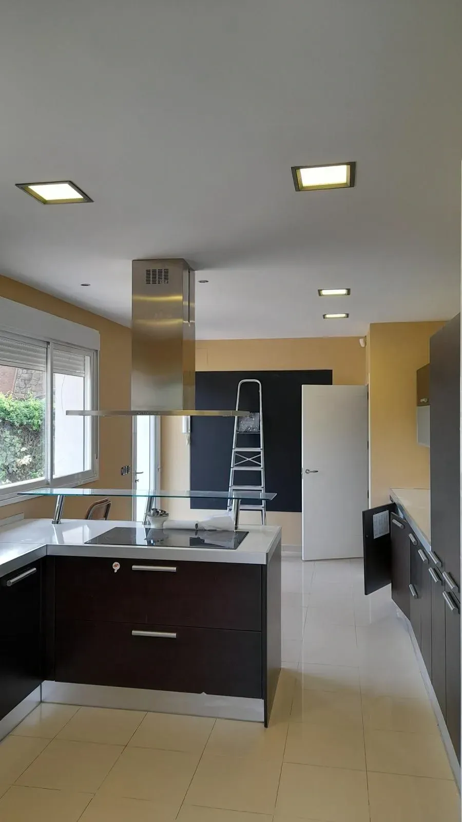 Cocina moderna con isla oscura, campana extractora de acero inoxidable, armarios blancos y suelo de baldosas beige.