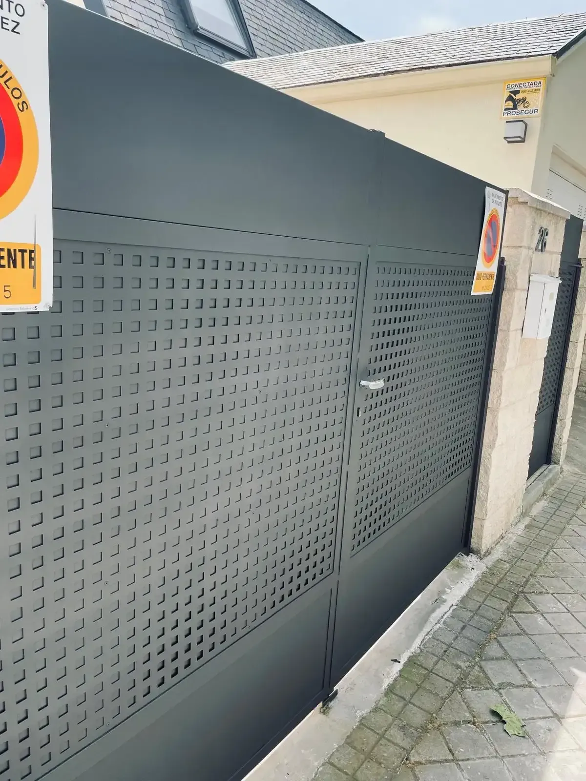 Puerta de metal perforado gris junto a una acera y un muro de estuco en el exterior.