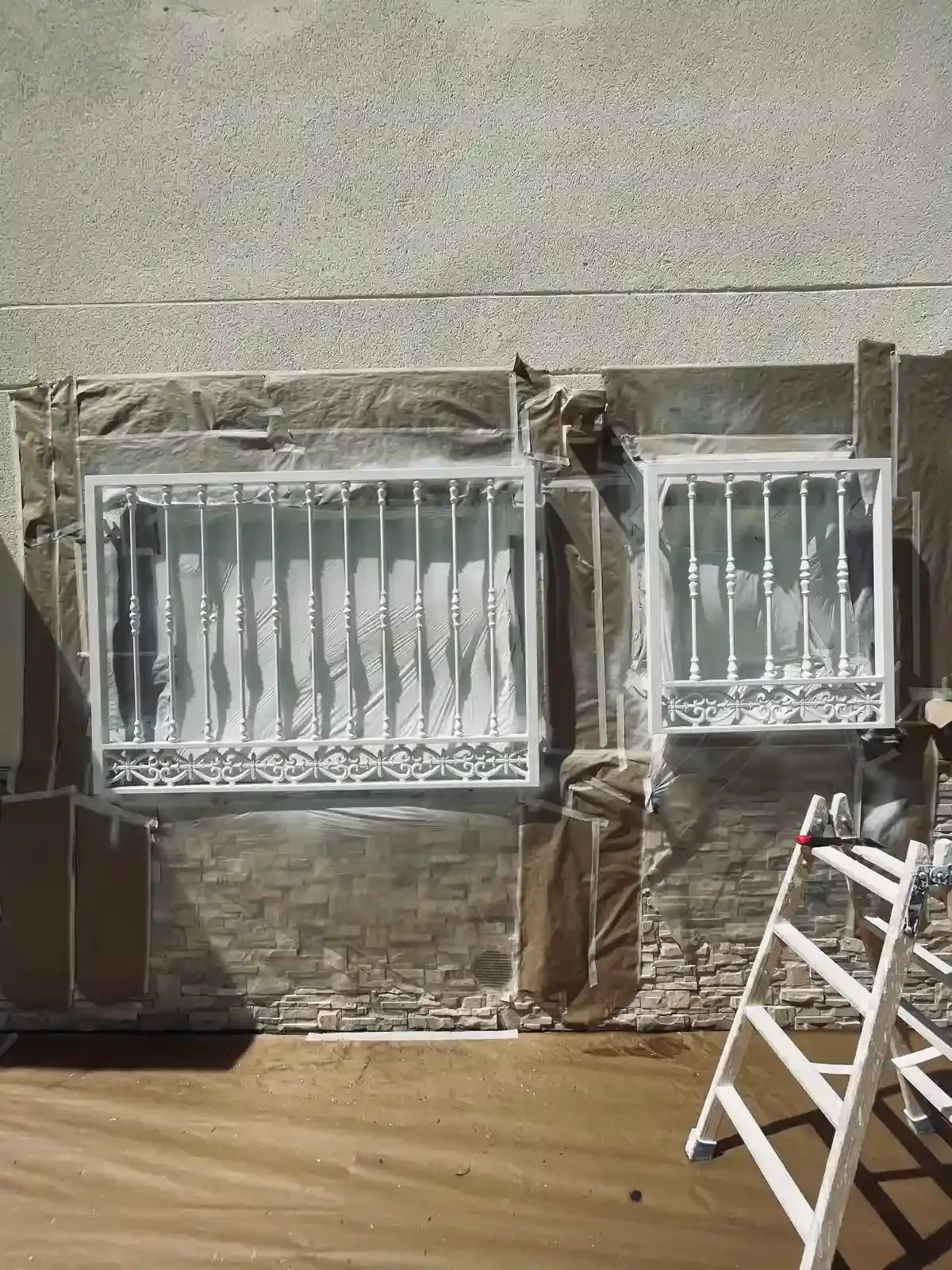 Dos ventanas con barrotes en una pared de estuco, con una cubierta protectora y una escalera apoyada cerca.
