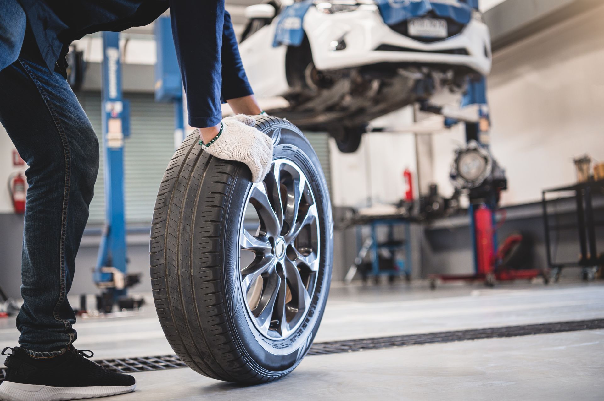 Ein Mechaniker in blauem Hemd und Jeans rollt in einer Autowerkstatt einen Autoreifen. Im Hintergrund ist ein Auto erhöht zu sehen.