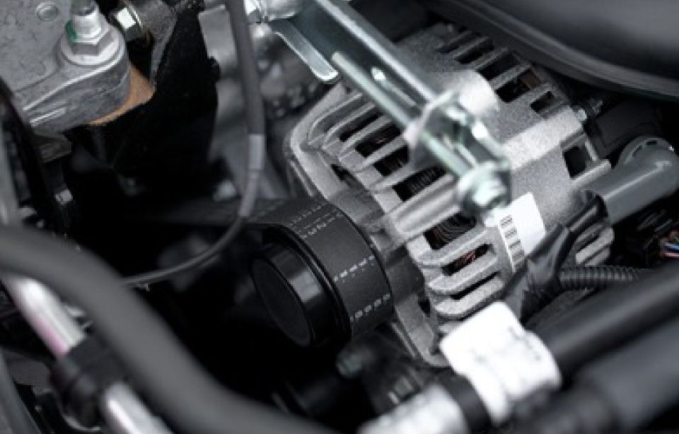 Ein Mechaniker in blauem Hemd und Jeans rollt in einer Autowerkstatt einen Autoreifen. Im Hintergrund ist ein Auto erhöht zu sehen.