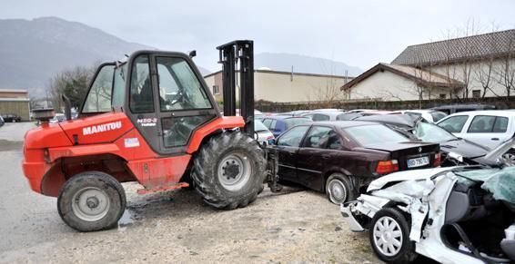 Un chariot élévateur rouge remorque une voiture endommagée sur un parking, par temps nuageux.