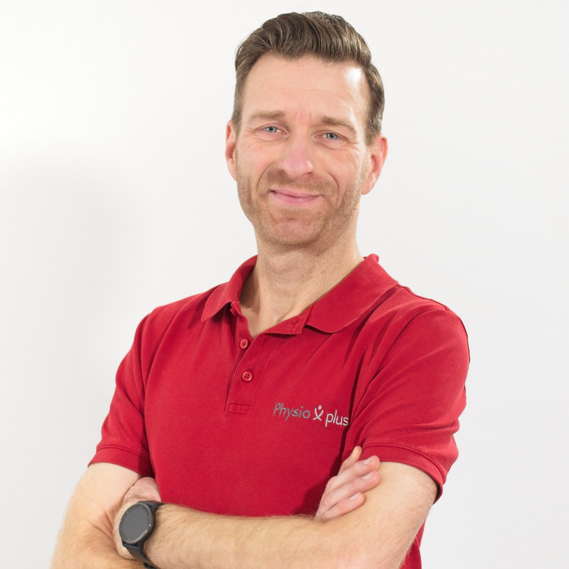 Ein Mann in einem roten Poloshirt steht mit verschränkten Armen vor einer weißen Wand.
