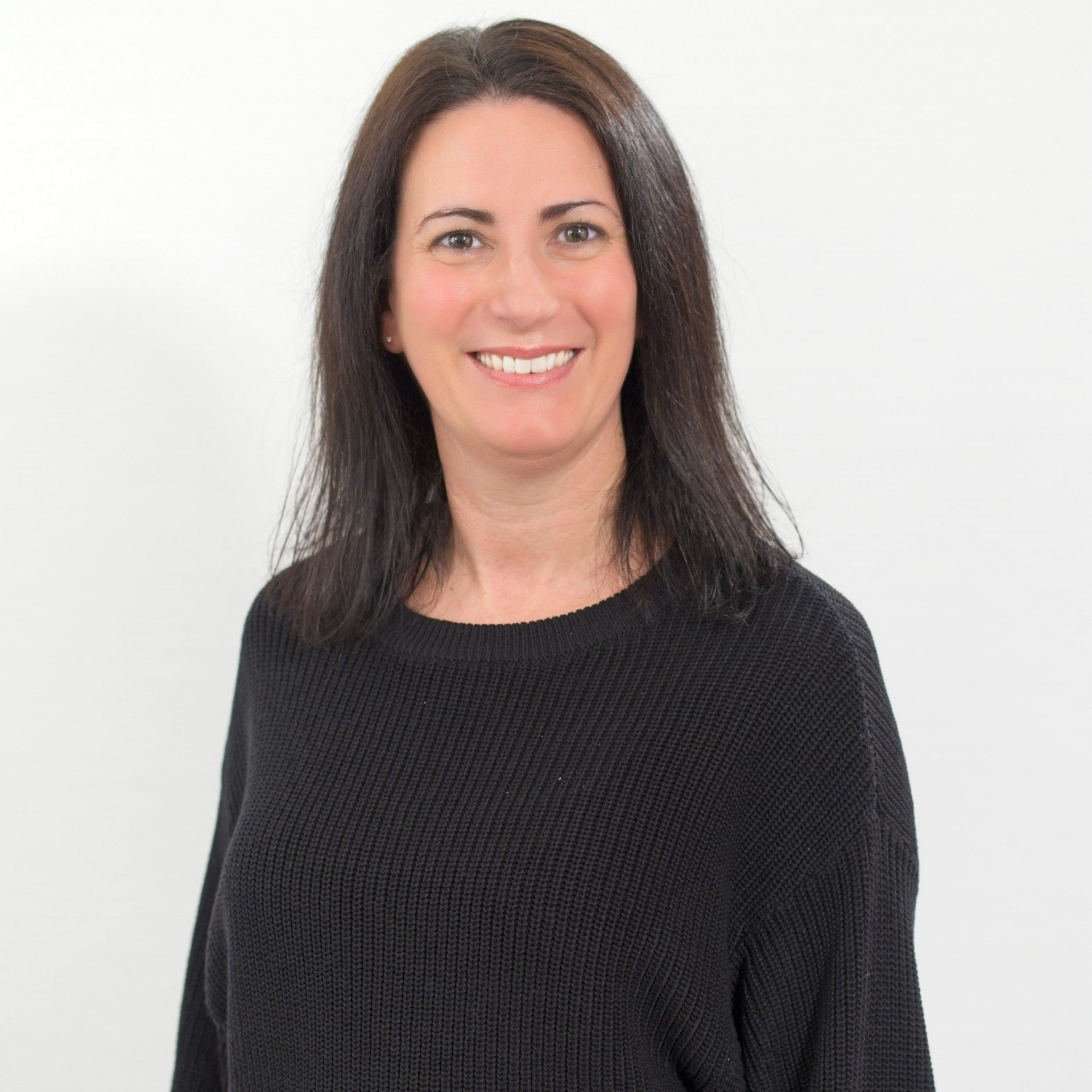 Eine Frau in einem schwarzen Hemd und Jeans steht vor einer weißen Wand.