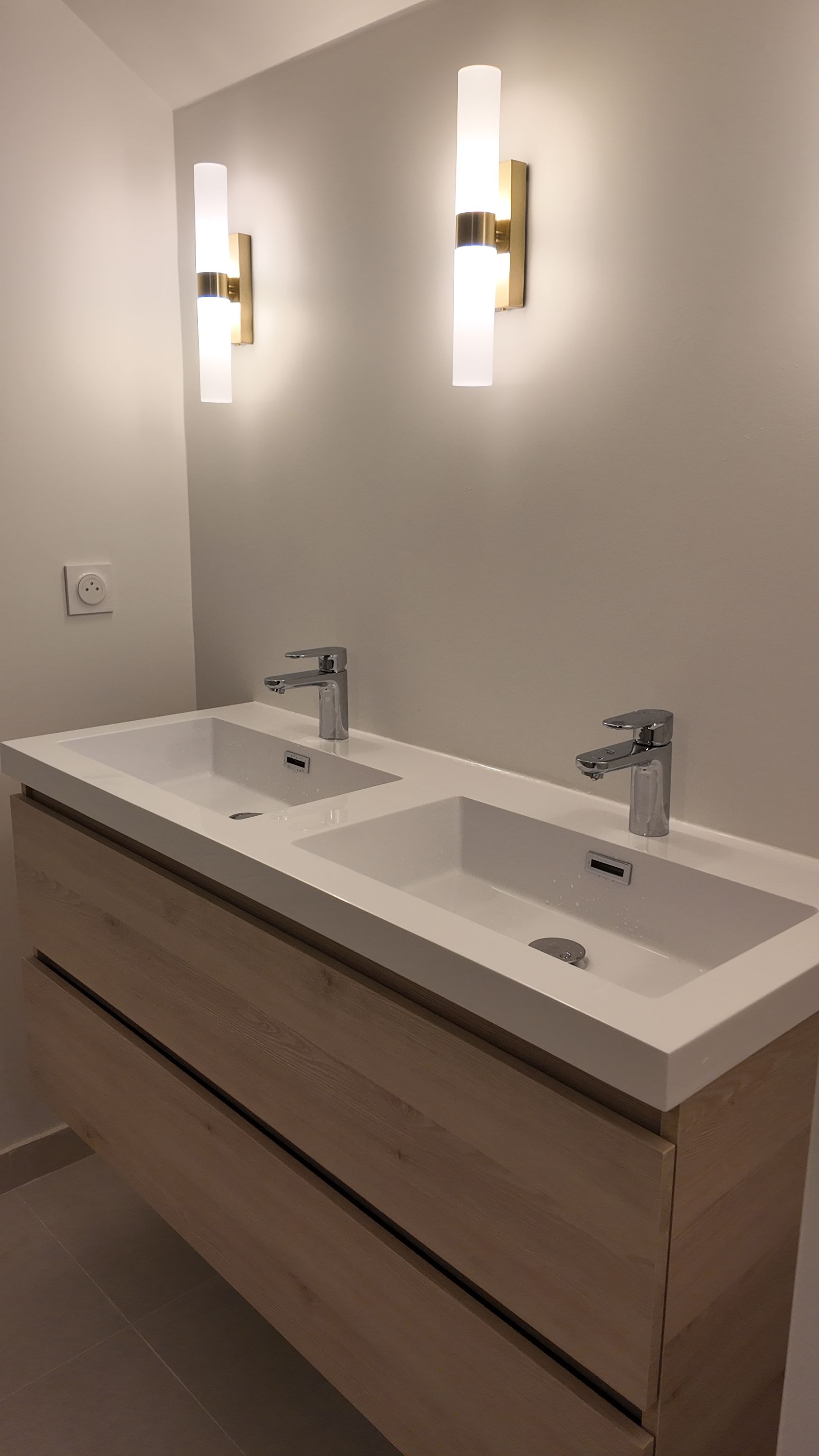 Meuble-lavabo double vasque avec robinetterie chromée, deux appliques murales sur un mur blanc. Armoire en bois.