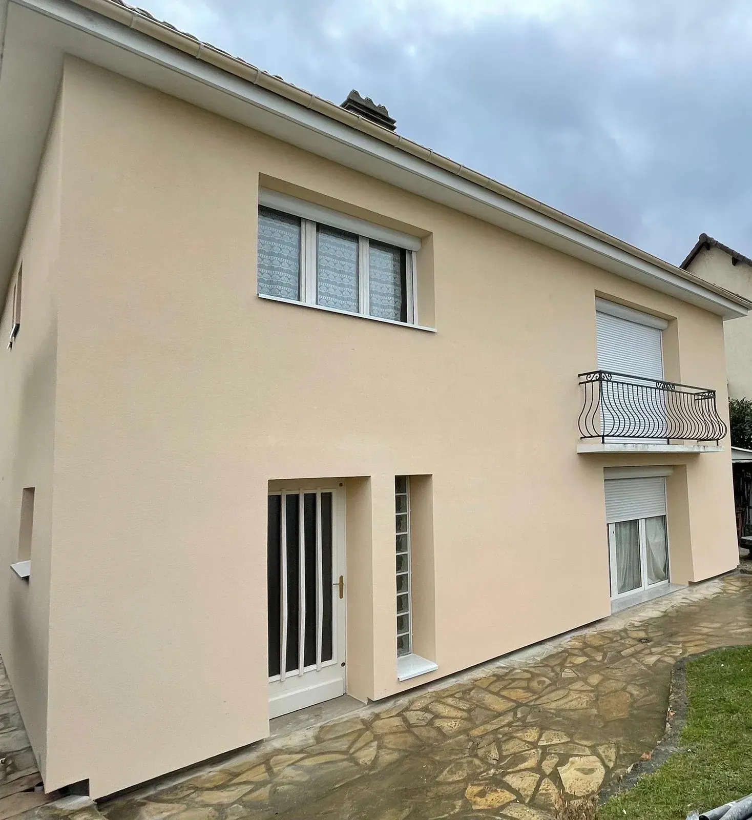 Maison à deux étages avec un enduit beige, des fenêtres et des boiseries blanches, un petit balcon avec une rambarde en fer forgé et un ciel nuageux.