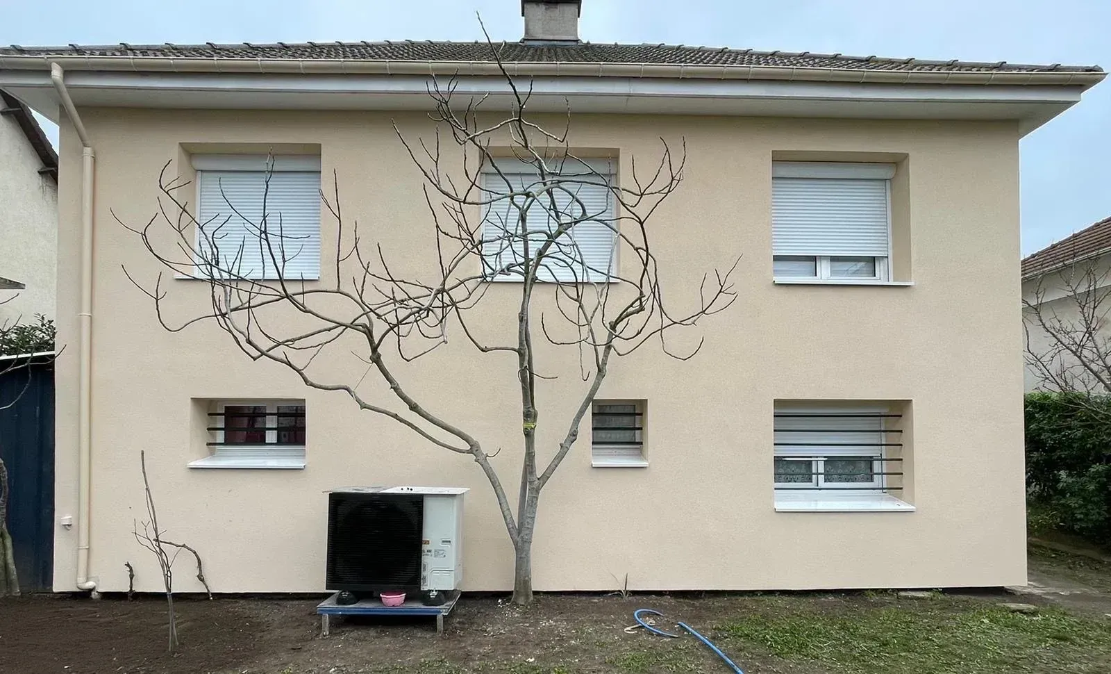 Maison beige à deux étages avec stores blancs fermés, un arbre sans feuilles et un climatiseur.