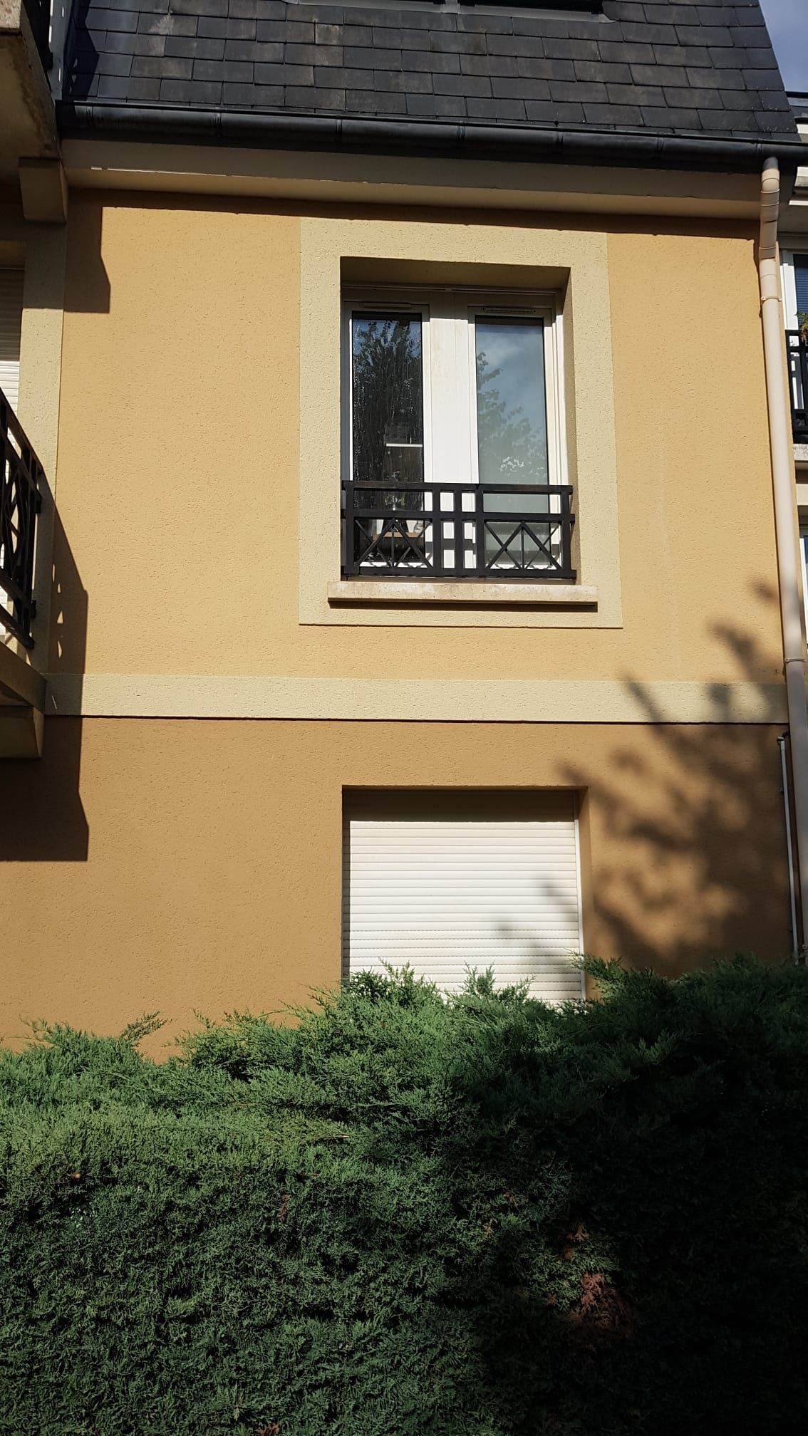 Façade jaune d'un bâtiment avec fenêtre, balcon et volet blanc fermé au-dessus de la verdure.