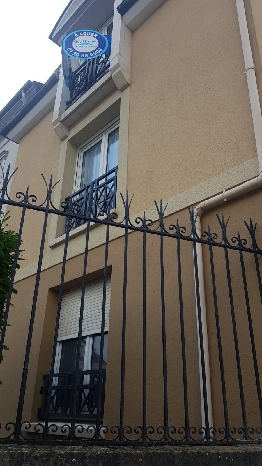 Immeuble beige avec grille en fer forgé. Balcon, fenêtres. Panneau au-dessus du balcon.