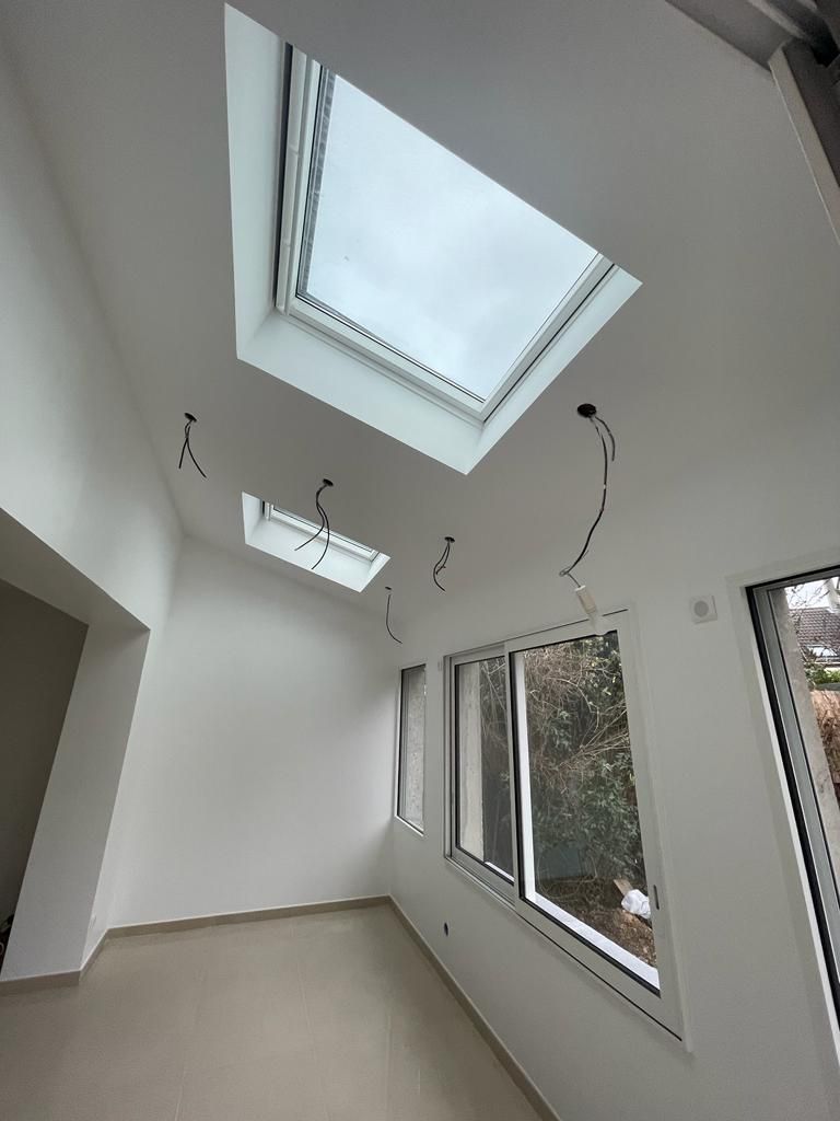 Pièce intérieure avec deux puits de lumière, une fenêtre, des murs blancs et des fils électriques apparents.
