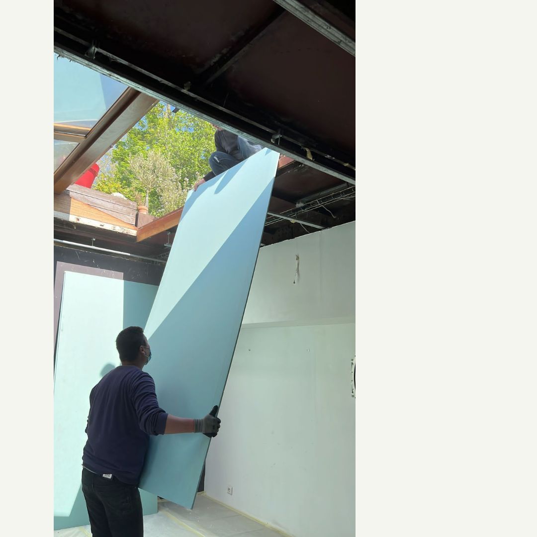 Deux ouvriers installent un grand panneau isolant bleu à l'intérieur d'un bâtiment.