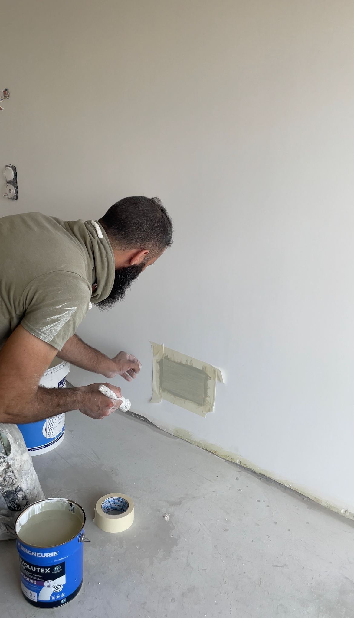 Un homme applique de l'enduit sur un mur, masquant la zone près du sol avec du ruban adhésif. Un pot de peinture se trouve à proximité.