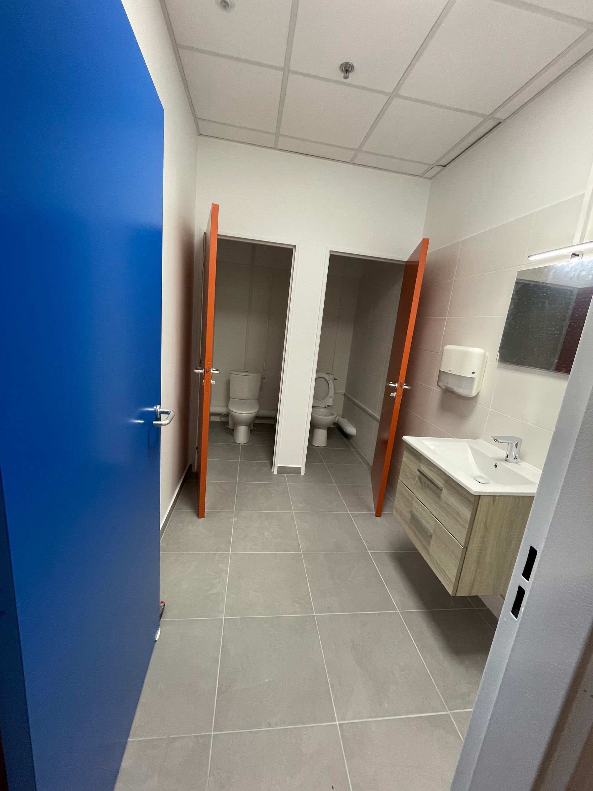 Salle de bains avec deux cabines de toilettes, un lavabo et une porte bleue sur la gauche.