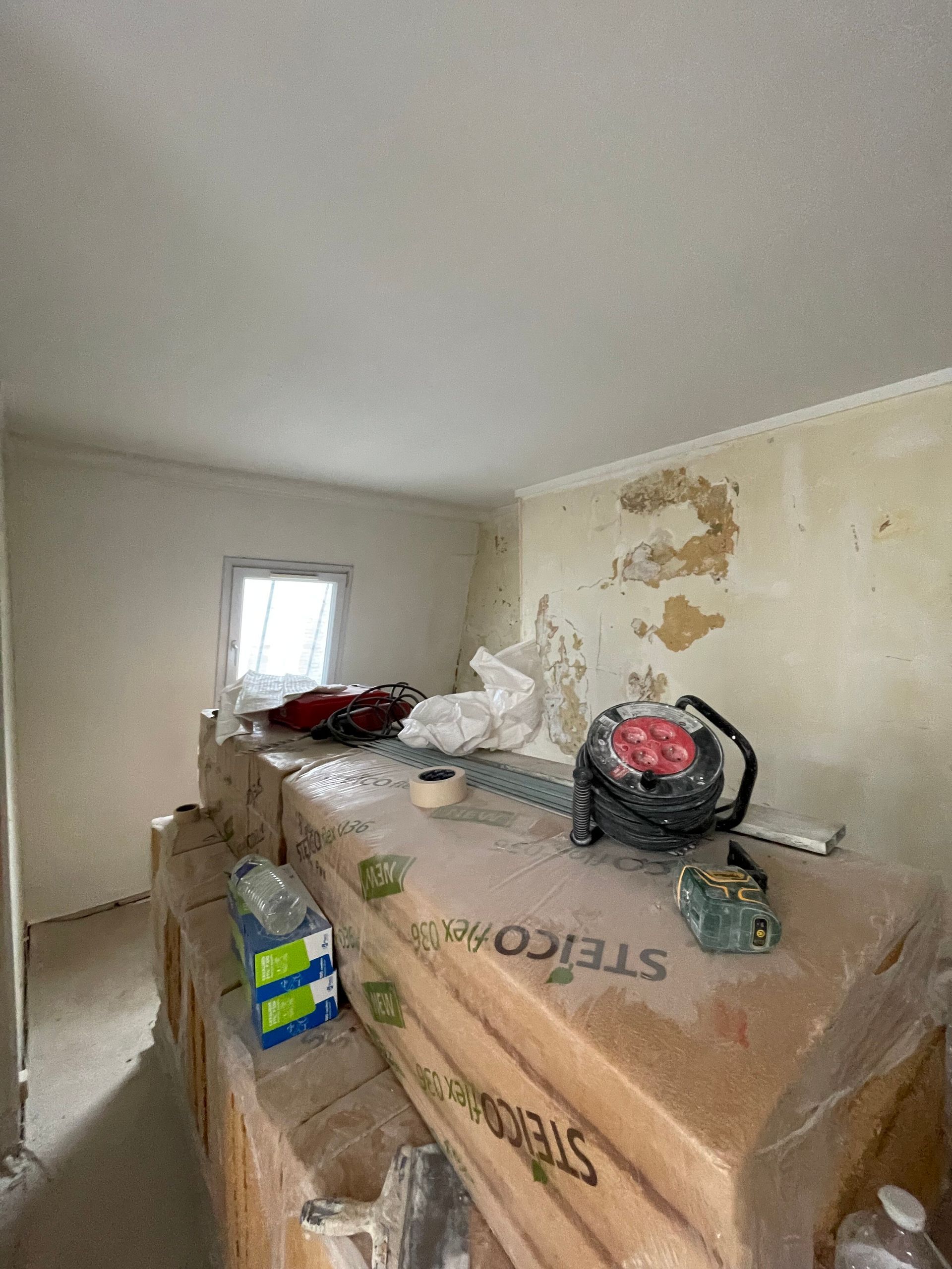 Pièce en construction avec isolation, outils et matériaux. Fenêtre visible.