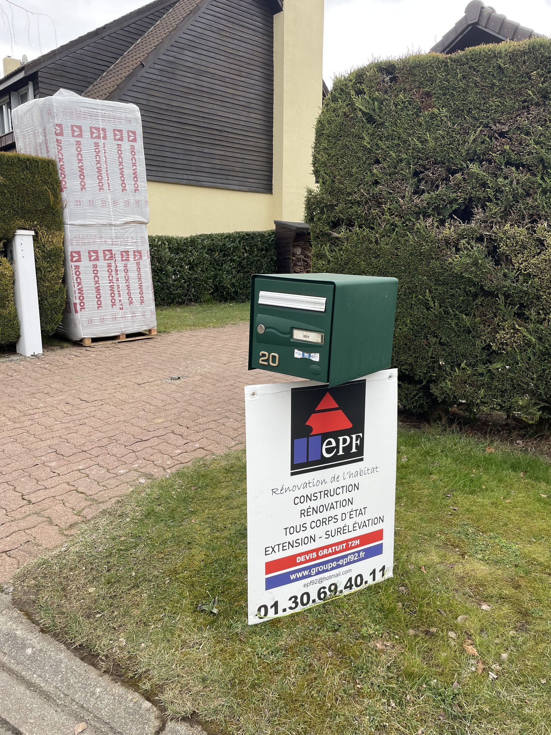 Boîte aux lettres verte sur un panneau, sur la pelouse ; maison en arrière-plan.