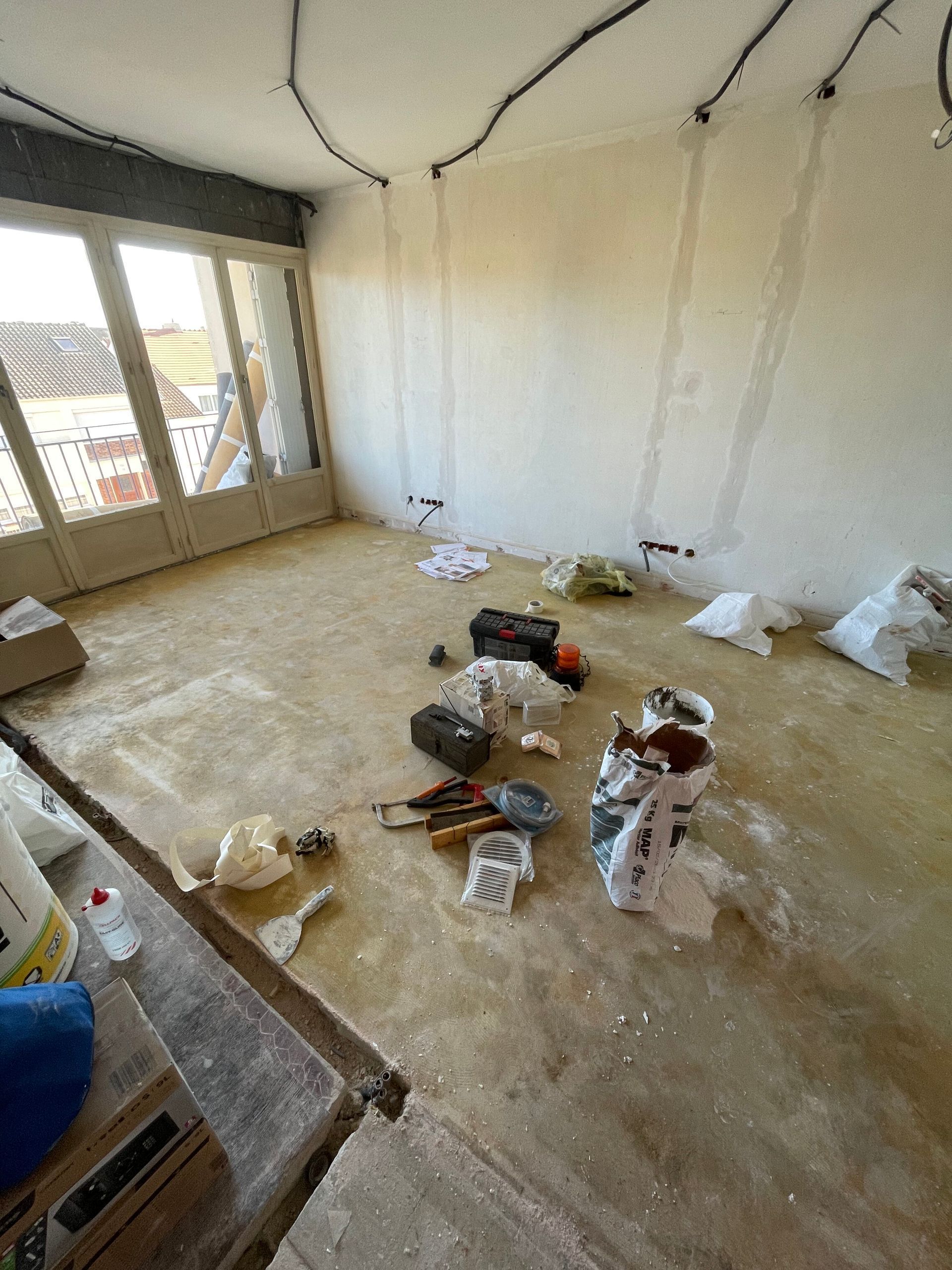 Pièce en travaux, sol poussiéreux, outils, câbles électriques, sacs de matériaux, une porte donnant sur un balcon.