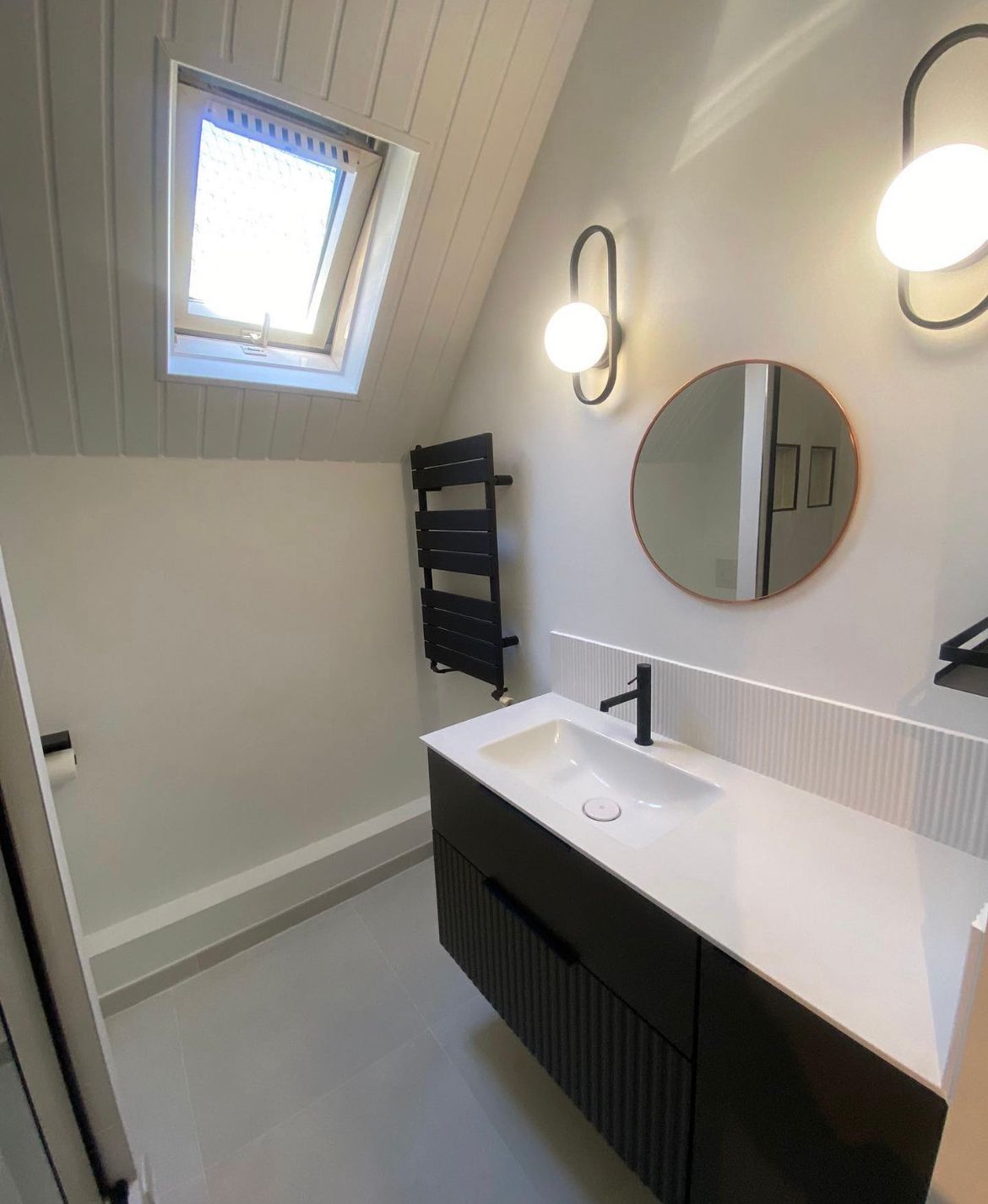 Salle de bains avec puits de lumière, meuble-lavabo noir, miroir rond et deux appliques murales.