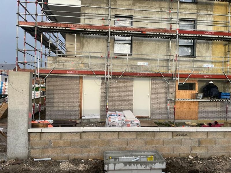 Bâtiment en construction, des échafaudages entourent la structure. Isolation visible, portes et fenêtres présentes.