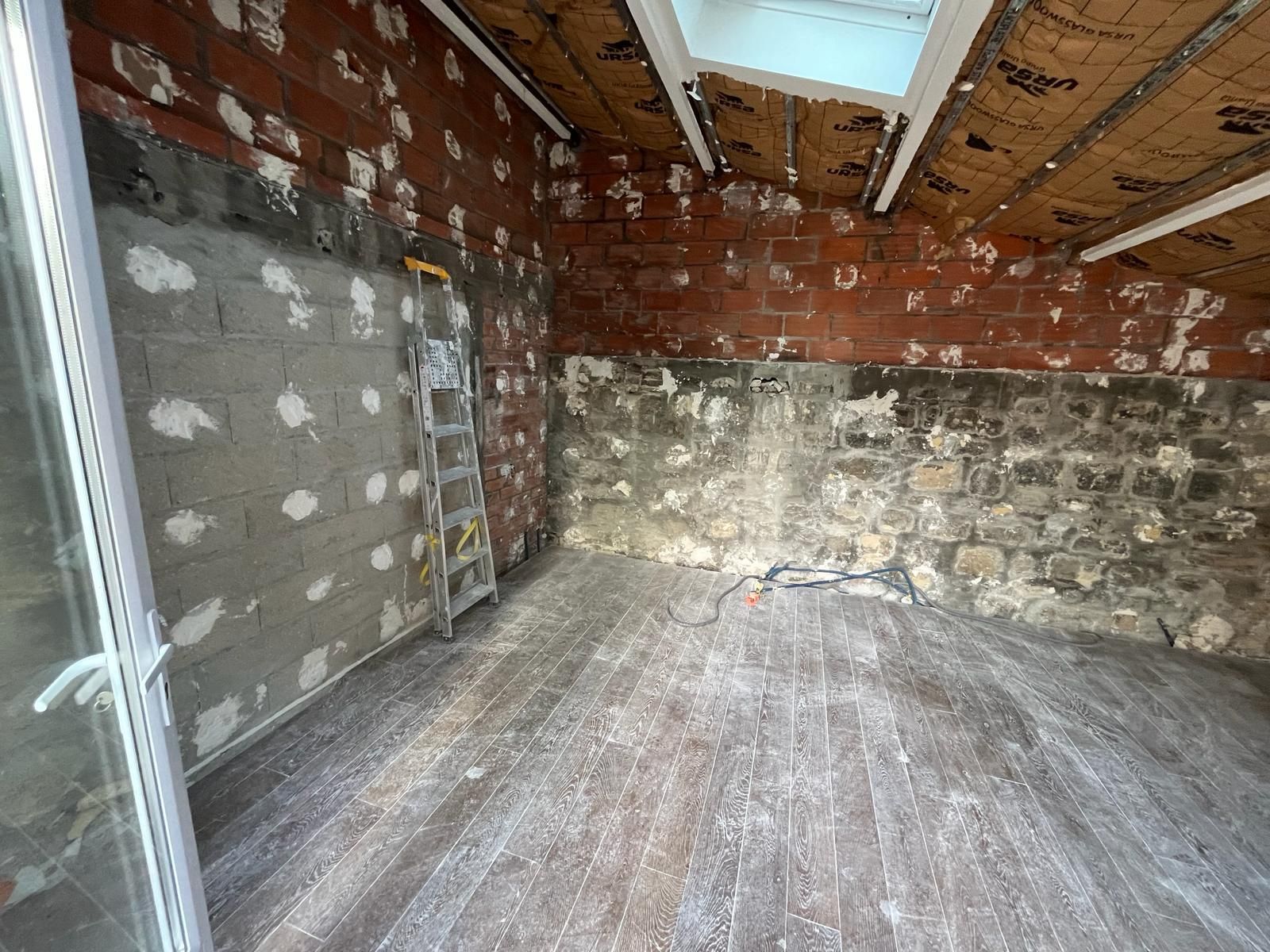 Pièce en rénovation avec murs en briques apparentes, murs en pierre et parquet ; une échelle est appuyée contre un mur.