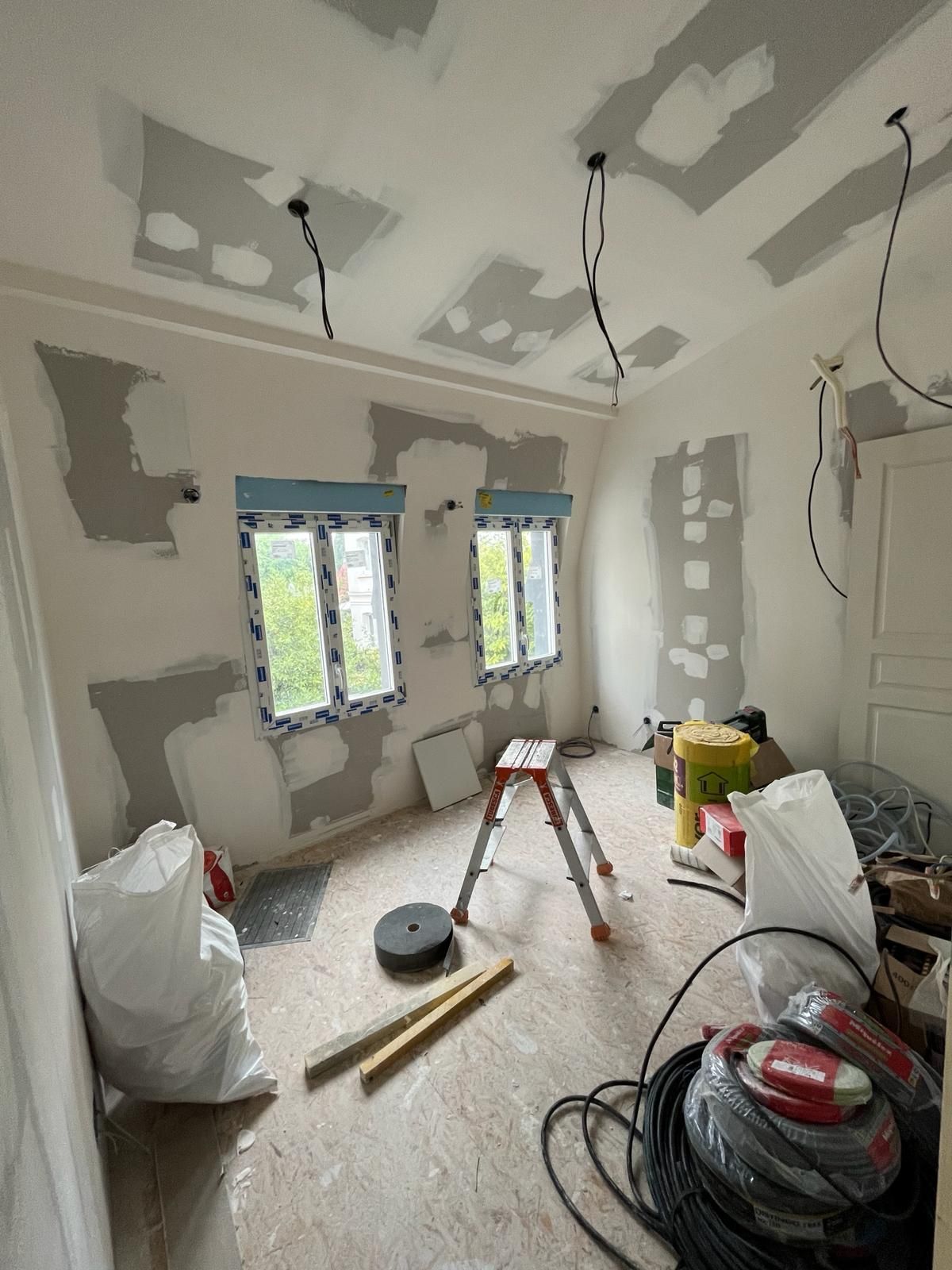 Pièce en rénovation ; cloisons sèches, câbles électriques apparents et matériaux de construction éparpillés. Deux fenêtres et un escabeau sont visibles.