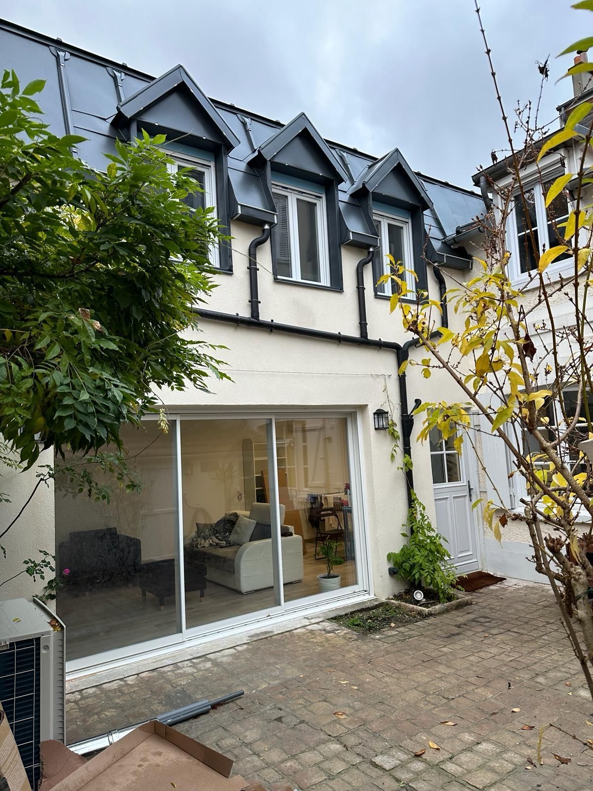 Un bâtiment de deux étages aux murs blancs, au toit gris et doté de trois lucarnes, avec de grandes portes coulissantes en verre et un patio en briques.