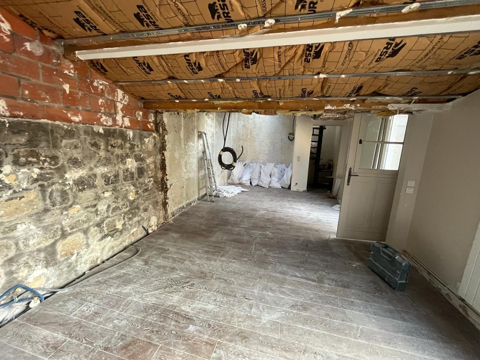 Rénovation intérieure en cours ; briques, pierres et bois apparents ; plafond et sol non finis ; porte visible.