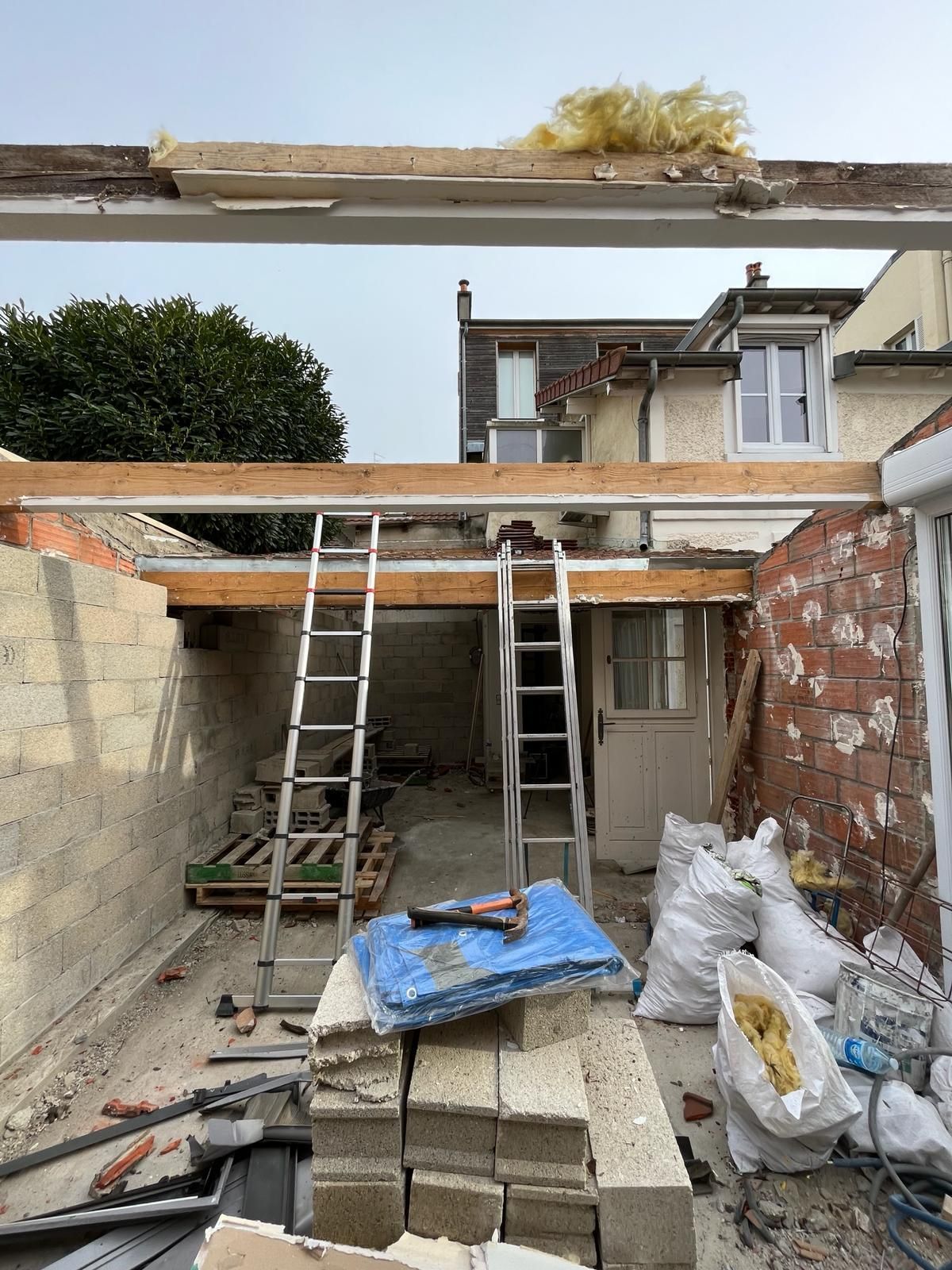 Chantier de construction avec deux échelles, poutres apparentes et débris dans un jardin.