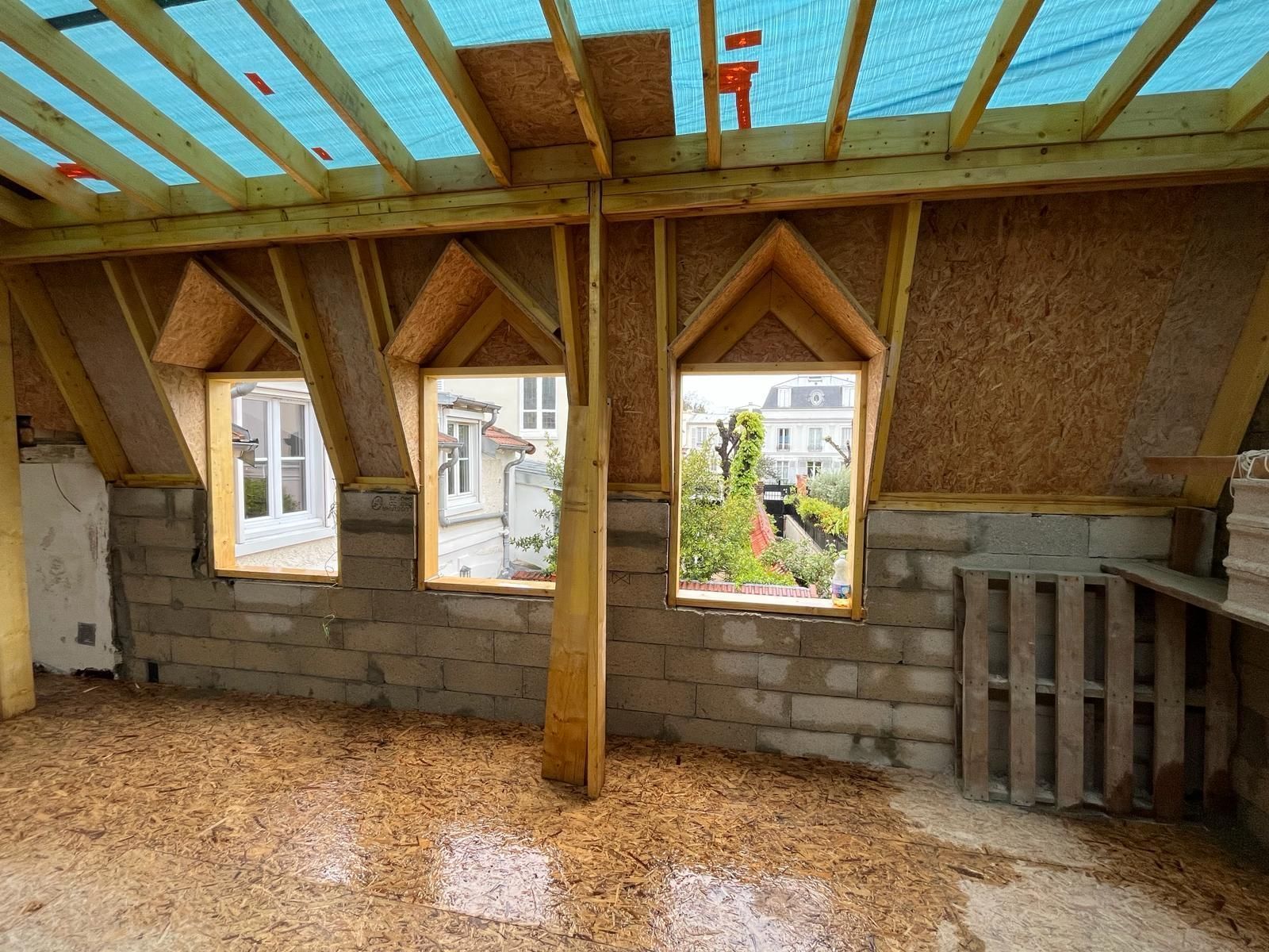 Vue intérieure d'un bâtiment en construction, montrant des fenêtres, des poutres en bois et des murs en briques apparentes.