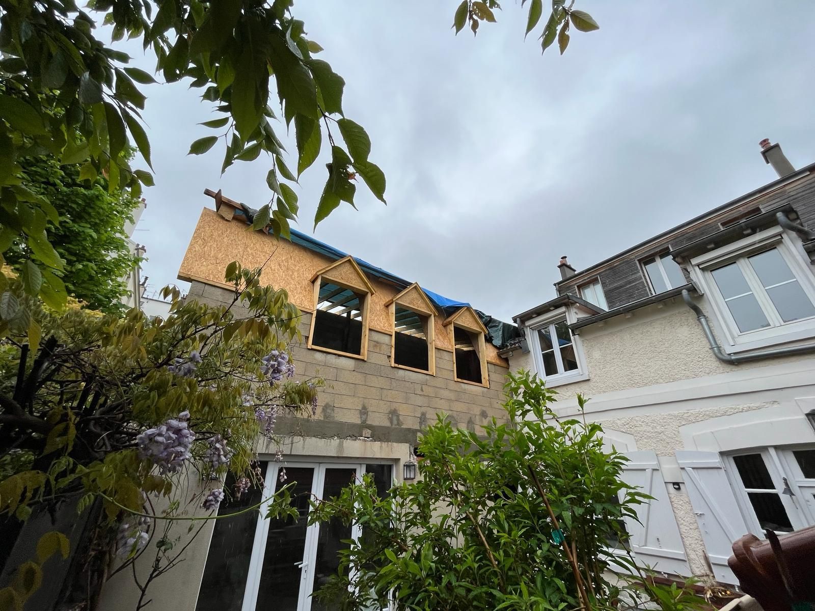 Maison en construction ; nouvelles lucarnes en cours d’aménagement, charpente en bois apparente. Ciel couvert, verdure au premier plan.
