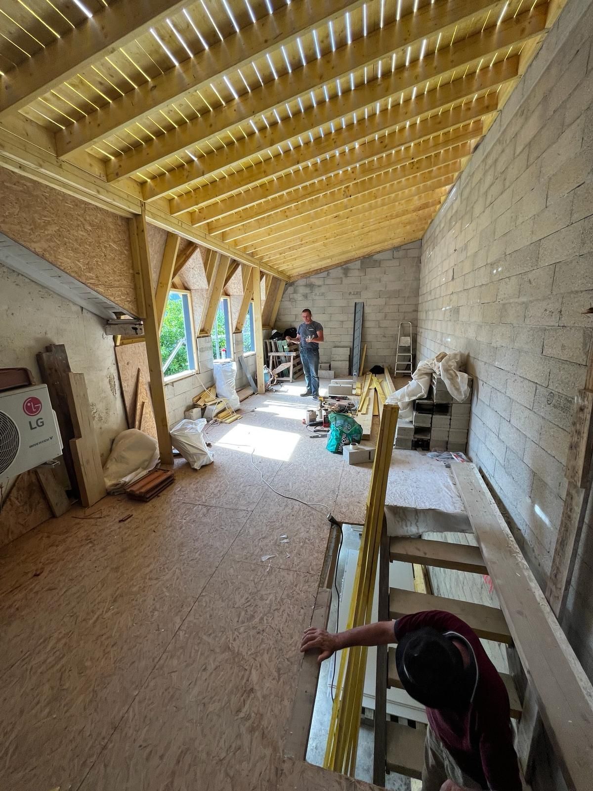 Intérieur d'une pièce en construction avec des poutres en bois apparentes et une personne debout au centre.