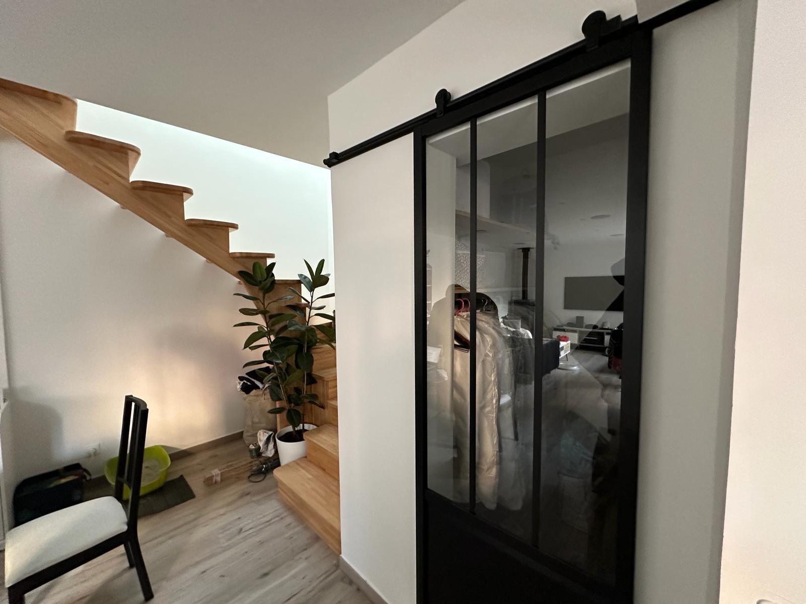 Porte coulissante en verre à cadre noir, montée sur un rail, dévoilant une pièce aux murs blancs, un escalier et une chaise.