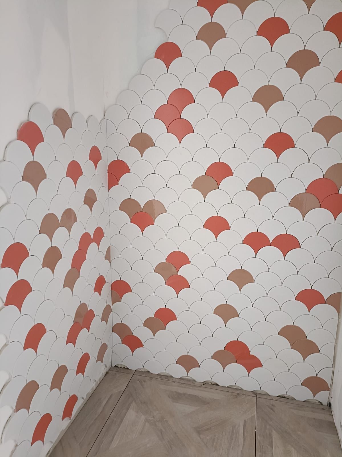 Un mur est recouvert de carreaux blancs, corail et beiges en forme d'éventail. Un coin de la pièce est parqueté.