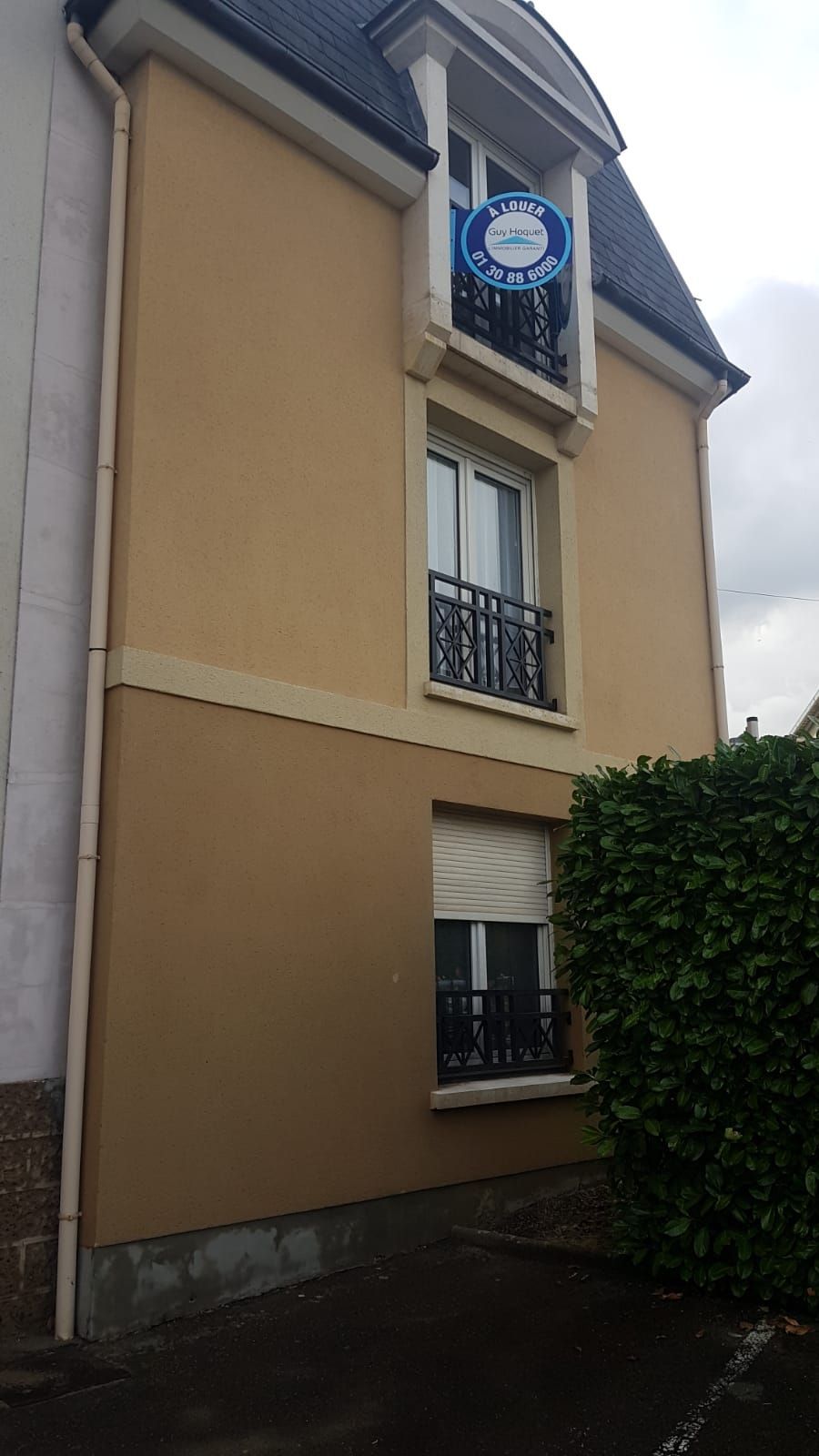Immeuble beige de trois étages avec toit noir ; fenêtres avec balcons. Panneau « À vendre » visible.