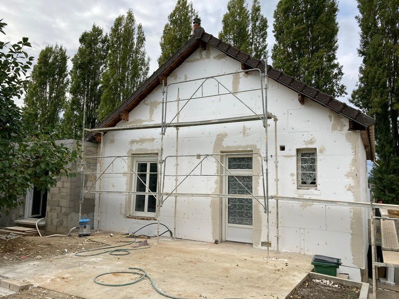 Maison en construction, avec échafaudages. Extérieur blanc, toit marron. Arbres en arrière-plan, ciel nuageux.