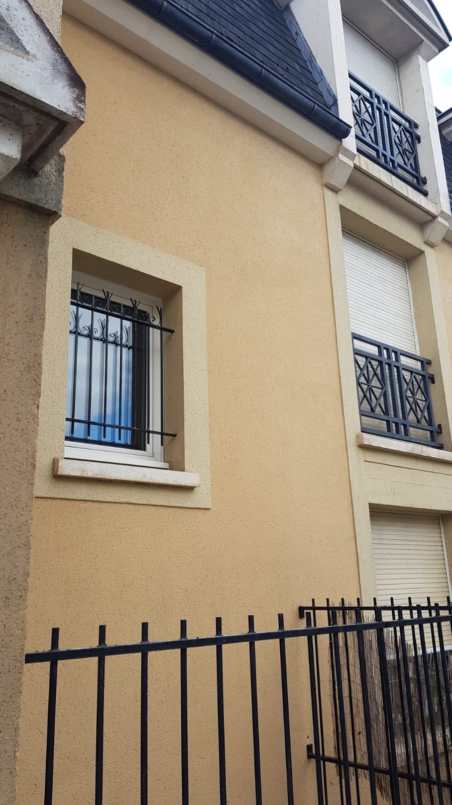 Bâtiment jaune avec une fenêtre munie de barreaux de sécurité. Une rambarde noire se trouve au premier plan.