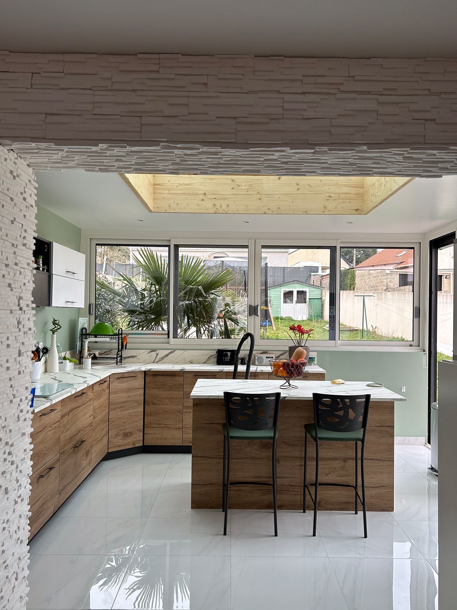 Cuisine moderne avec placards en bois, îlot central avec tabourets et grandes fenêtres donnant sur un jardin.