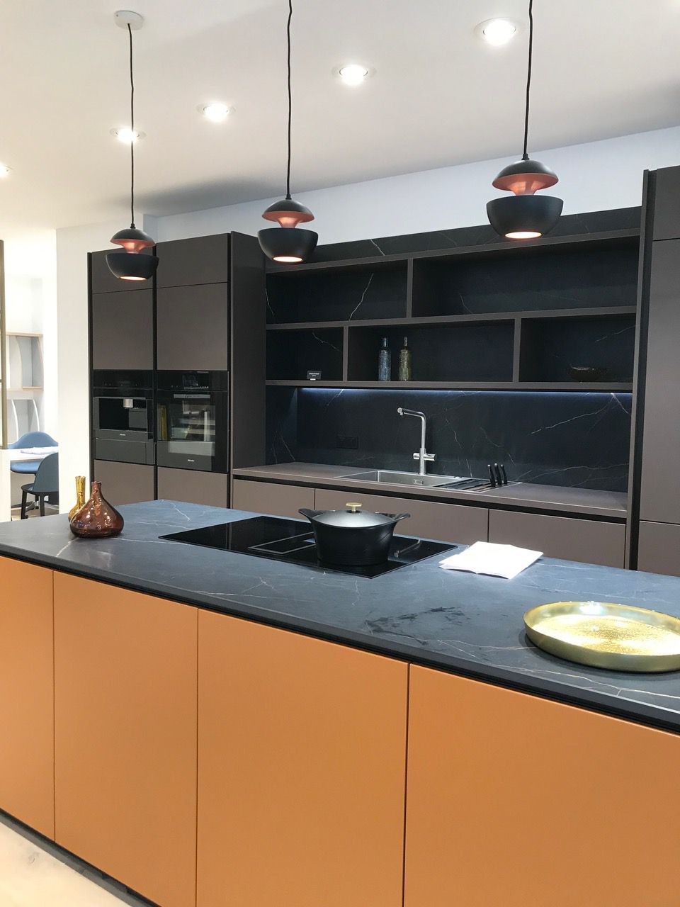 Cuisine moderne avec îlot central orange, placards gris foncé, trois suspensions lumineuses et plaque de cuisson intégrée.
