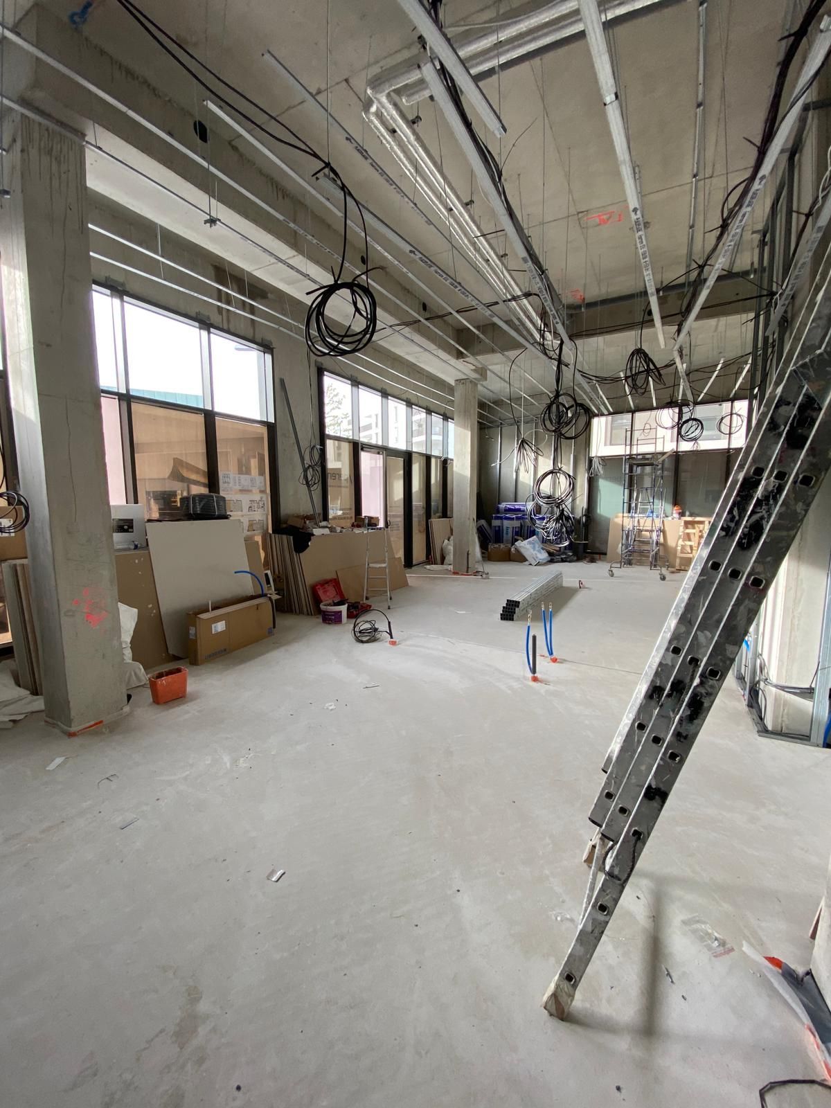 Intérieur d'un bâtiment en construction, avec des sols en béton, des câbles électriques apparents et une échelle appuyée contre un mur.