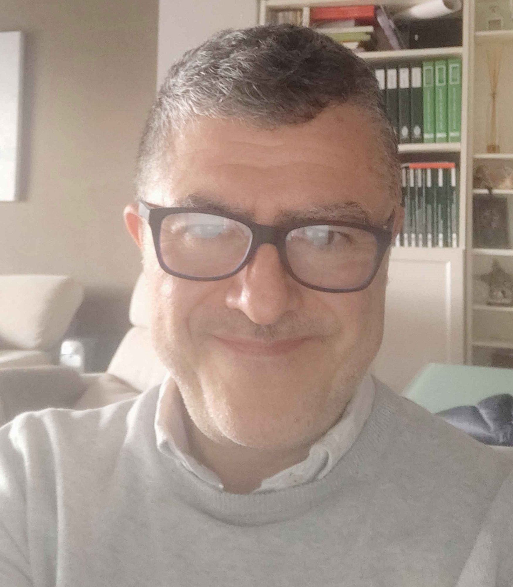 Hombre con gafas, sonriente, delante de una estantería, vistiendo un suéter gris claro.