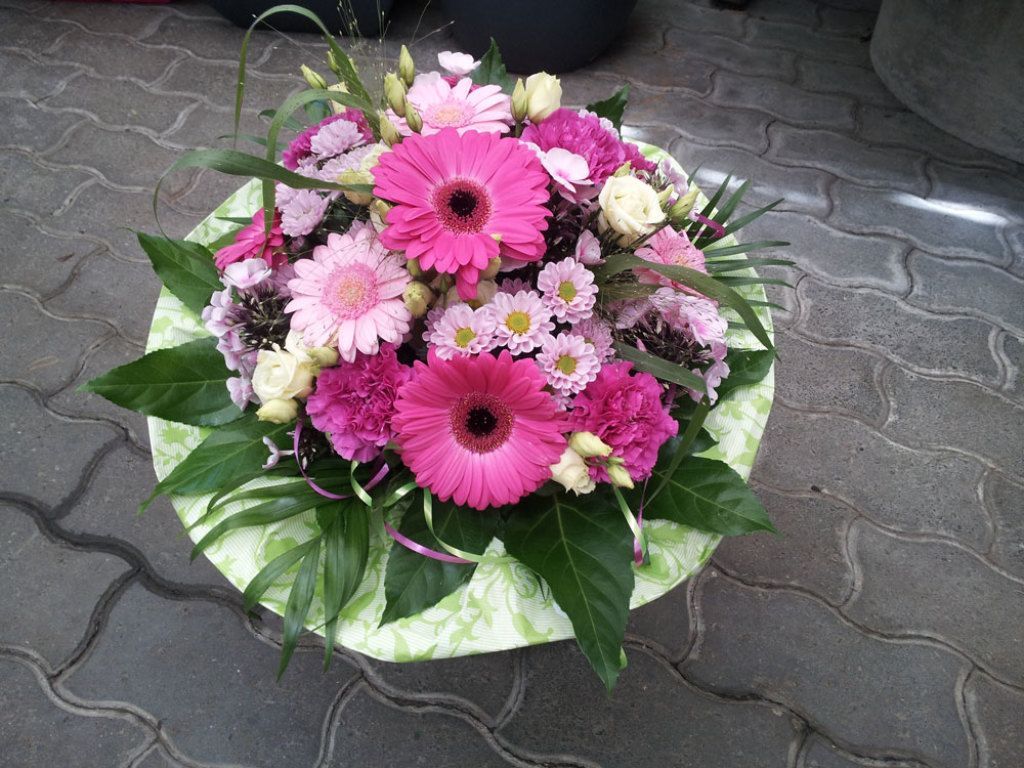 Blumenarrangement mit rosa Gerbera, Nelken und Grünpflanzen in einem runden Behälter auf einer Ziegeloberfläche.
