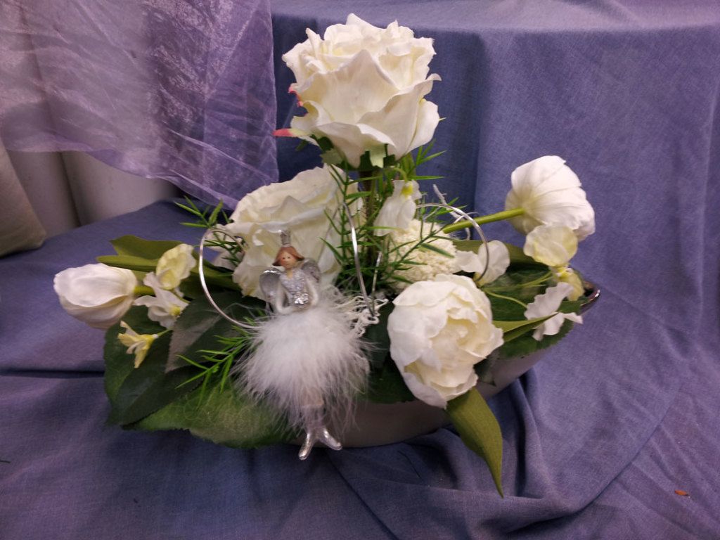 Weißes Blumenarrangement in einer silbernen Schale mit einer kleinen Feenfigur auf einer lila drapierten Oberfläche.