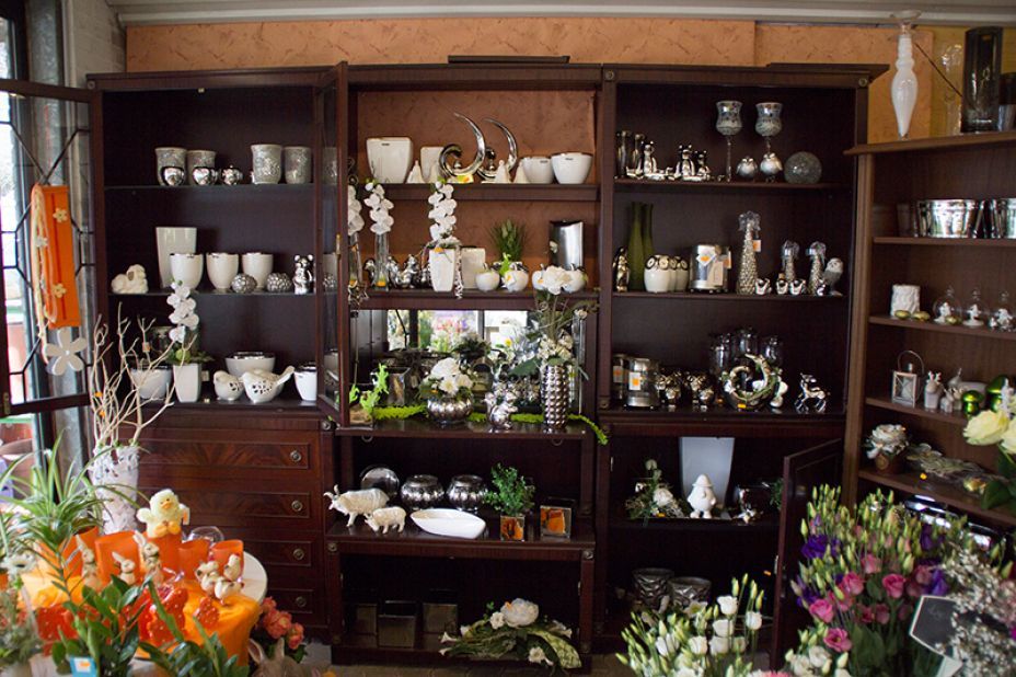 Ausstellungsregale in einem Geschäft mit weißen und silbernen Dekorationsartikeln, Blumen und Holzmöbeln.