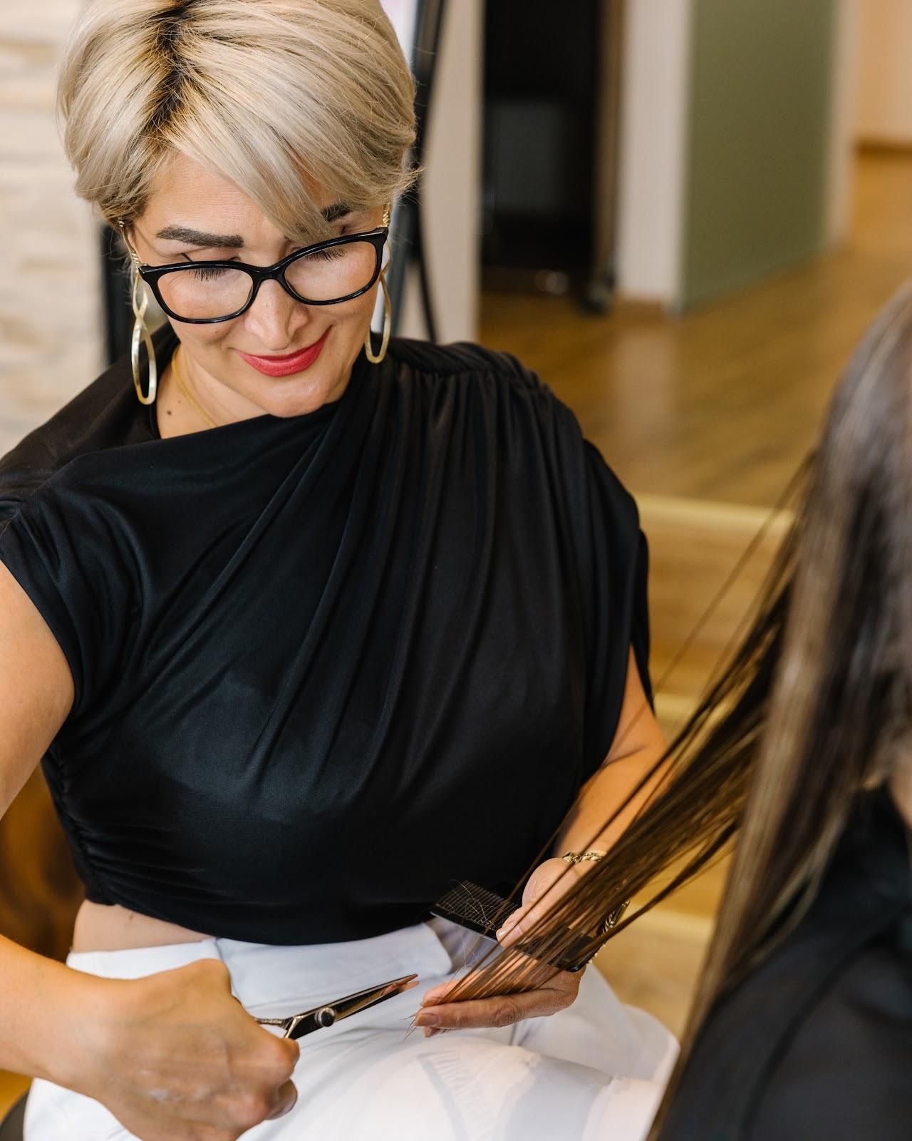 Friseurin schneidet Kundin mit der Schere die Haare. Blondes Haar, schwarzes Oberteil, Brille. Helle Umgebung.