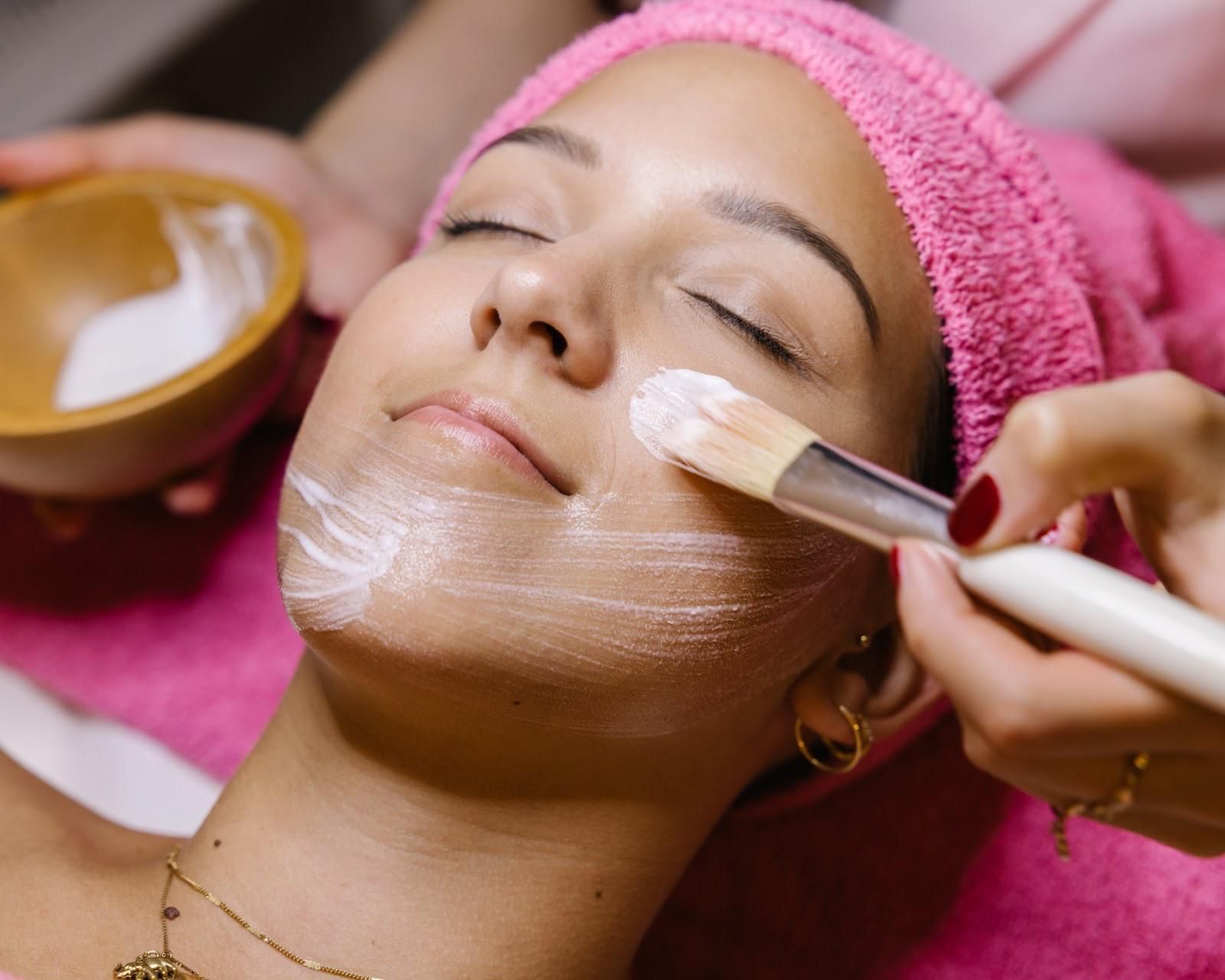 Person bei der Gesichtsbehandlung; Creme wird mit einem Pinsel aufgetragen. Rosa Handtuch, Holzschale.