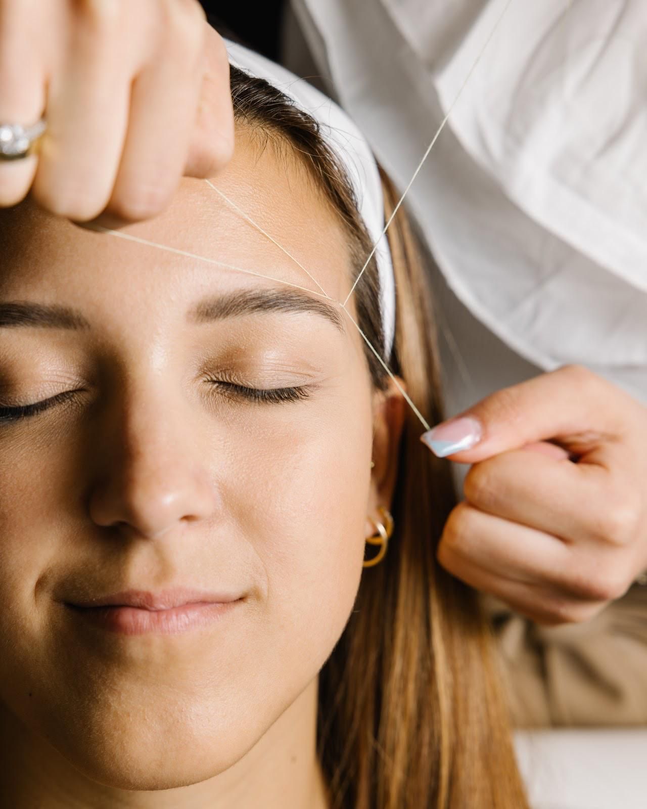 Frau lässt sich die Augenbrauen zupfen. Person hält Faden in der Nähe der Augenbraue, Augen geschlossen.