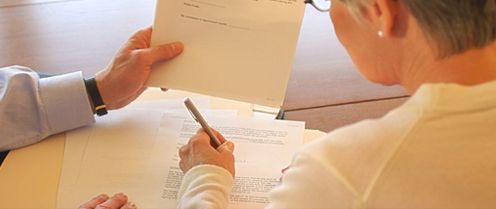 Dos personas revisan documentos en un escritorio; una sostiene una carpeta y la otra escribe con un bolígrafo.