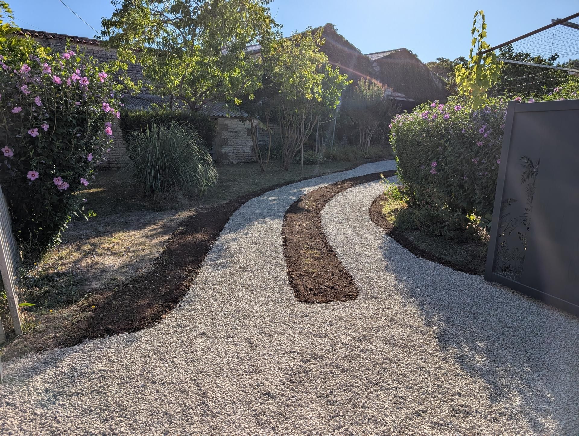 Allée de gravier avec un chemin de terre serpentant entre des buissons et un portail, sous un ciel ensoleillé.