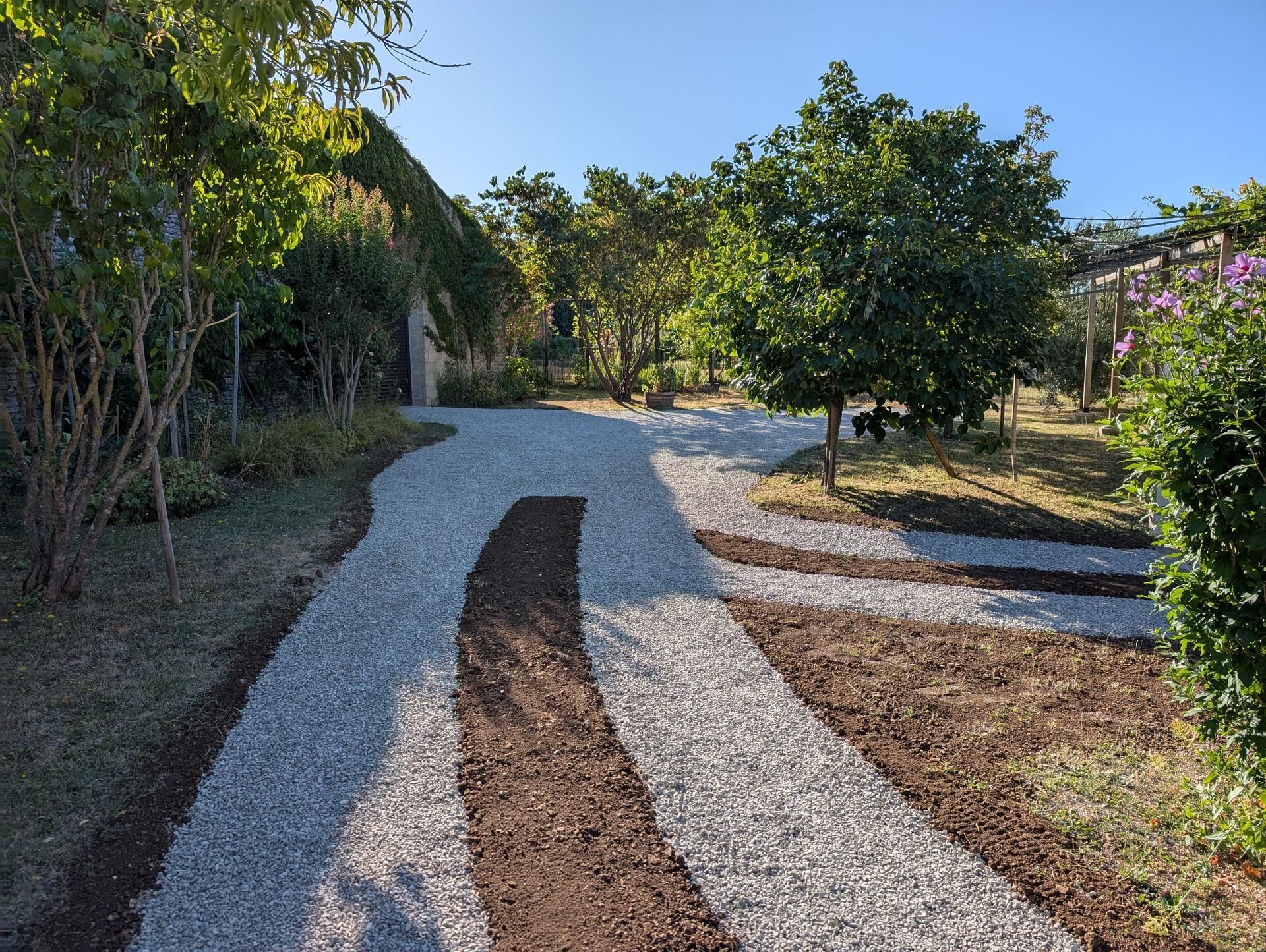 Allée de gravier serpentant à travers un jardin luxuriant avec des arbres, des plantes et du soleil.
